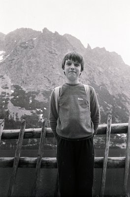 Young individual in mid-20th-century outdoor attire—sweater, pants, and backpack—standing on a wooden railing near a frozen a...