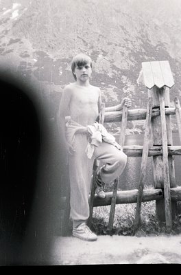 Young boy in mid-20th-century outdoor setting, shirtless with rolled-up pants, leaning on rustic wooden fence. Blurred foregr...