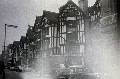 Black-and-white street scene featuring Tudor-style timber-framed buildings with exposed beams and decorative brickwork, likel...