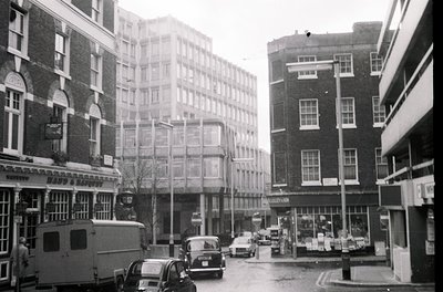 Mid-century urban street scene featuring brick facades, vintage cars, and a bus. Prominent signage reads "Lloyds Bank" and "S...