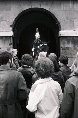 Black-and-white photo of a public event featuring a mounted police officer addressing a crowd through an archway. Uniform inc...