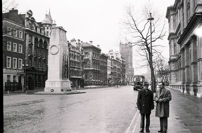 Two men in 1950s-era suits stand near Nelson’s Column in London’s Trafalgar Square. Surrounding architecture features ornate ...