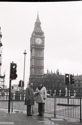 Two men in 1960s-era overcoats pose near Big Ben, London. Urban scene with traffic lights and railings. Classic black-and-whi...