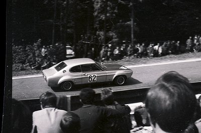 Vintage race car () in motion on a tree-lined road, likely a 1960s rally event. Crowd watches from roadside, suggesting publi...