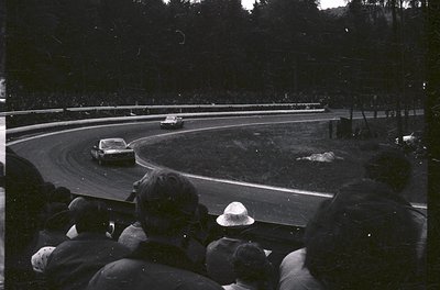 Black-and-white race scene: spectators in hats watch a vintage sports car navigate a sharp curve on a wet track, surrounded b...