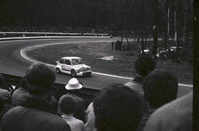 Classic 1960s rally car navigating a sharp curve on a tree-lined track, surrounded by spectators. The compact vehicle feature...