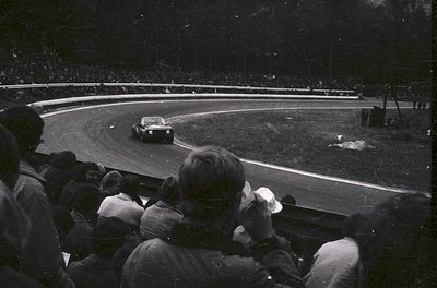 Black-and-white shot of a vintage race car navigating a sharp curve on a wet track, surrounded by spectators in grandstands. ...