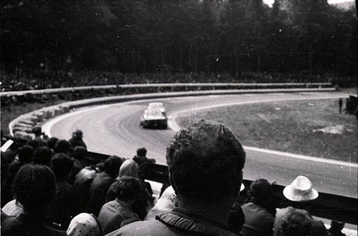 Black-and-white shot of a vintage race car navigating a banked oval track, surrounded by spectators. Mid-20th century racing ...