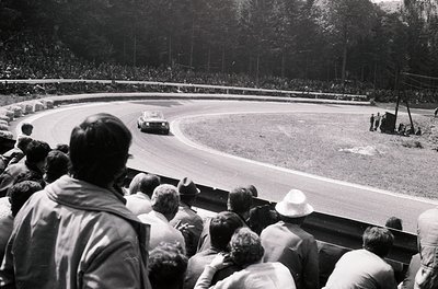 Classic 1960s motorsport scene: spectators packed into grandstands watch a vintage race car navigate a sharp turn on a tree-l...