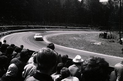 Classic race scene: vintage sports car navigating a sharp curve on a tree-lined track, surrounded by spectators. Mid-20th cen...