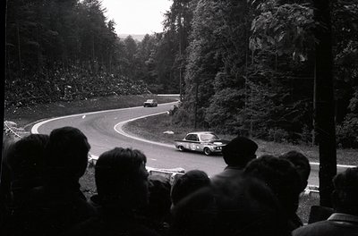 Classic black-and-white shot of a **rally race** on a winding mountain road, featuring vintage cars navigating tight curves. ...