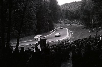 Vintage race car navigating a winding forest road during a rally, surrounded by dense spectators. Black-and-white photo captu...
