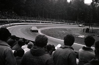 Black-and-white shot of a vintage race car navigating a sharp left curve on a tree-lined track, surrounded by dense crowds. M...