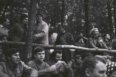 Group of men in military-style uniforms and caps gathered outdoors in a forested area, likely Eastern Bloc 1960s–70s. Uniform...