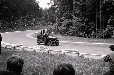 Vintage open-top race car navigating a winding road, surrounded by spectators on concrete barriers. Early 20th-century automo...