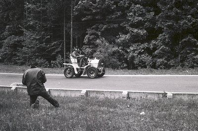 Vintage open-top vintage car racing on a rural road, with a lone figure in mid-stride beside the guardrail. Black-and-white s...