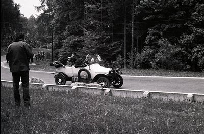 Vintage open-top race car on a rural road, surrounded by dense forest. Spectator in dark coat watches from guardrail. Mid-20t...