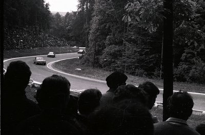 Black-and-white shot of a vintage race car navigating a winding mountain road, surrounded by spectators on the left. Classic ...