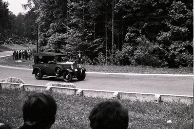Classic 1930s-era roadster with open-top design and side curtains, driving on a rural road flanked by dense forest. Crowd of ...