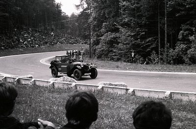 Vintage open-top race car navigating a winding road during a rally, surrounded by spectators. The car’s rounded fenders and s...