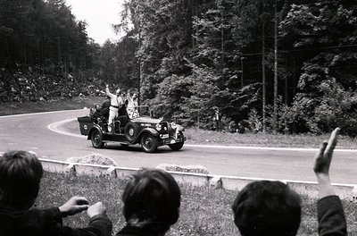 Vintage open-top car navigating a winding forest road, surrounded by onlookers waving. Classic 1930s–1940s roadster with visi...