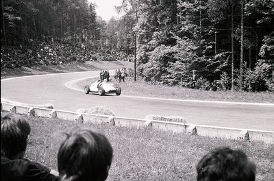 Classic open-wheel race car navigating a sharp curve on a wooded road, surrounded by spectators. Mid-20th century racing scen...