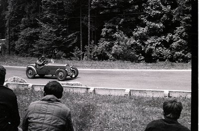 Vintage open-wheel race car navigating a curved road, surrounded by dense forest. Spectators in 1950s-era attire watch from g...