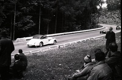 Classic race car navigating a winding forest road during a vintage rally, mid-20th century. Spectators in dark coats line the...