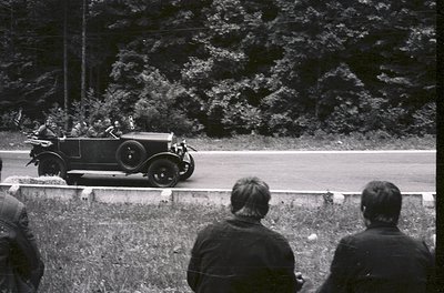 Classic 1930s roadster with open-top design and spoked wheels speeds past spectators on a rural road, framed by dense foliage...