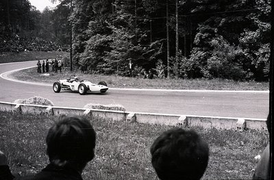 Open-wheel race car navigating a curved road during a vintage motorsport event, surrounded by spectators on grassy embankment...