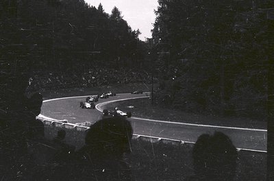 Vintage black-and-white shot of a winding mountain road with vintage race cars navigating a sharp curve. Dense forest and mis...