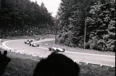 Classic 1960s Formula 1 race at Nürburgring, featuring vintage open-wheel cars navigating a sharp left-hand curve. Spectators...