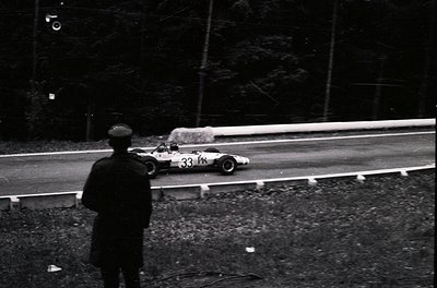 Spectator watches Formula 1 race car () on wet track, likely 1960s–1970s. Open-wheel design, high-speed cornering, and misty ...