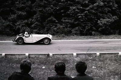 Classic 1930s–1940s roadster racing on a winding mountain road, likely Alpine region. Two spectators watch from embankment. L...