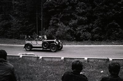 Vintage open-top touring car on a winding road, surrounded by dense forest. Spectators in foreground, likely at a rally or ev...
