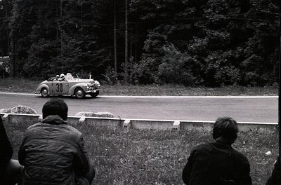 Vintage race car () navigating a winding forest road during a classic rally. Spectators in 1950s-era attire observe from gras...