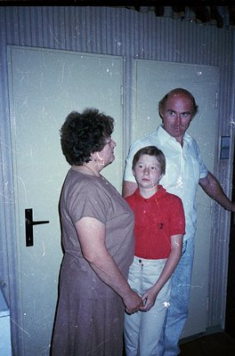 Indoor portrait of three individuals in a dimly lit hallway, likely mid-20th century. Woman in a dark dress stands left, hold...