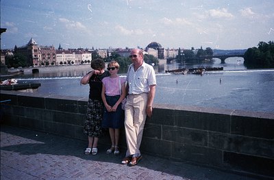 Three individuals pose on a stone bridge overlooking Prague’s Vltava River, 1960s. The man wears a light-colored suit, woman ...