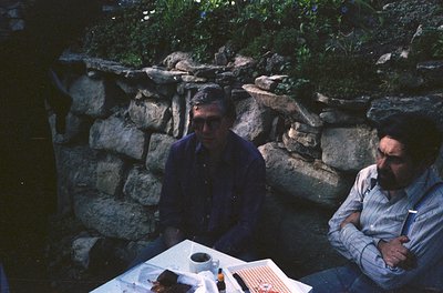 Two men seated outdoors at a stone-walled table, likely mid-20th century. Stone retaining wall with greenery behind them. Tab...