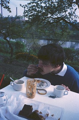Man eating outdoors by a body of water, mid-1970s. Table set with a half-eaten cake, coffee cup, and disposable cutlery. Indu...