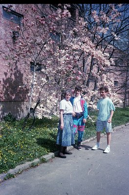 Three individuals pose outdoors beside blooming cherry blossoms and a brick building, likely 1980s–1990s USA. Woman in layere...