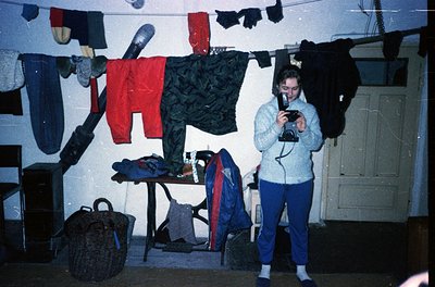 Vintage indoor laundry scene with 1970s-era clothing hung on lines. Person in a light sweater and blue pants holds a bulky ca...