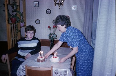 Vintage indoor birthday scene: woman in patterned blouse placing candles on two tiered cakes on a wooden table; teen boy seat...