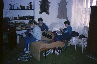 Three individuals study or work at a makeshift desk in a dimly lit room, likely a dormitory or shared living space. The table...