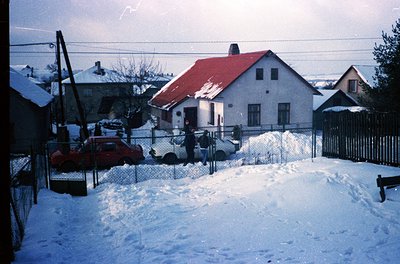 Snow-covered residential street with mid-century concrete houses, red-tiled roofs, and chain-link fences. Two individuals in ...