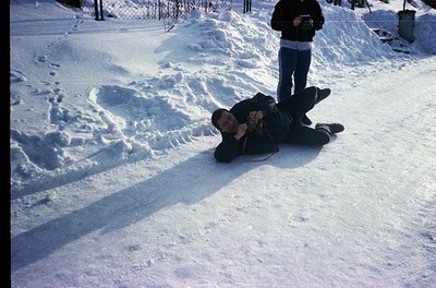 Vintage photo of two men in a snow-covered yard, likely mid-20th century. One man kneels in snow, holding a camera; the other...