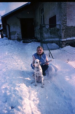 A person in winter attire sits on snow beside a Dalmatian, holding a book. Rustic wooden house with a metal fence and ladder ...