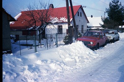 Snow-covered residential street with 1970s-era cars, likely Eastern Bloc region. Whitewashed houses with red-tiled roofs and ...