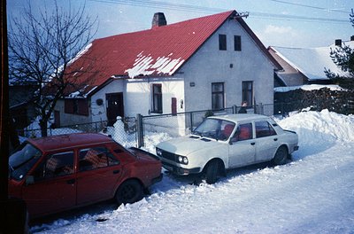 1970s-era residential home with red-tiled roof, snow-covered driveway, and two parked cars (maroon hatchback, white sedan). F...