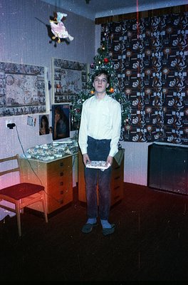 Young boy in 1970s-style sweater and rolled pants poses indoors, holding a wrapped gift. Surroundings include a vintage woode...